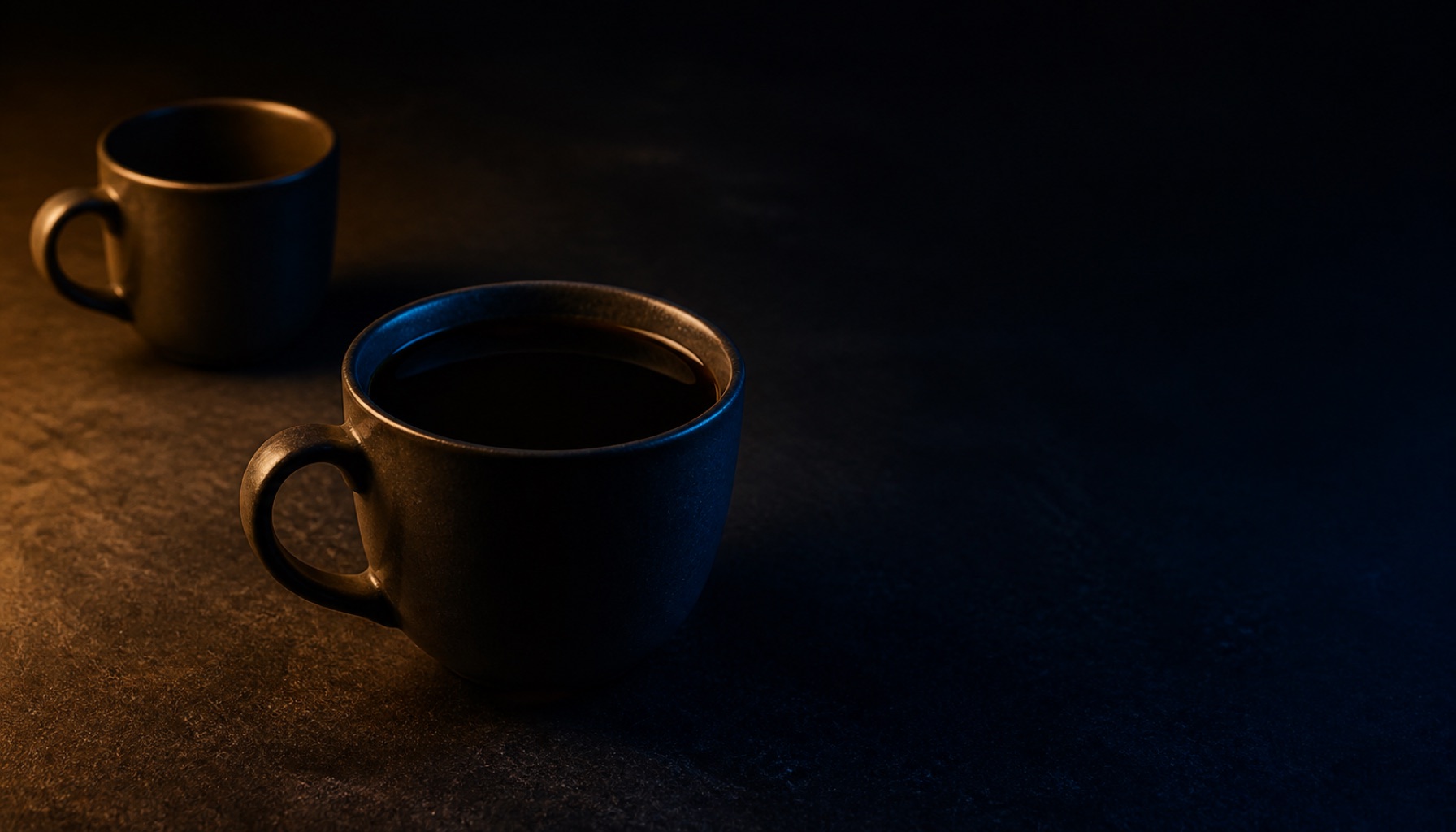 Two cups on a dark surface, one full and untouched in the foreground, the other empty and pushed away in shadow